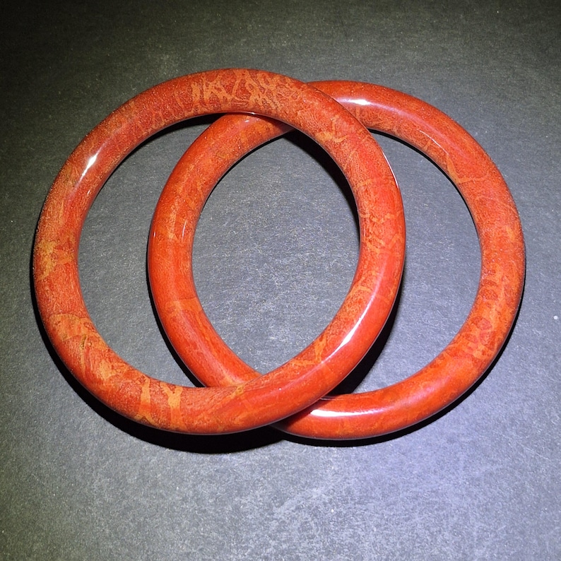 May include: Two red-orange bangles with a speckled pattern are interlocked. The circular bracelets have a glossy finish and appear to be made of a solid material. The background is a dark gray.