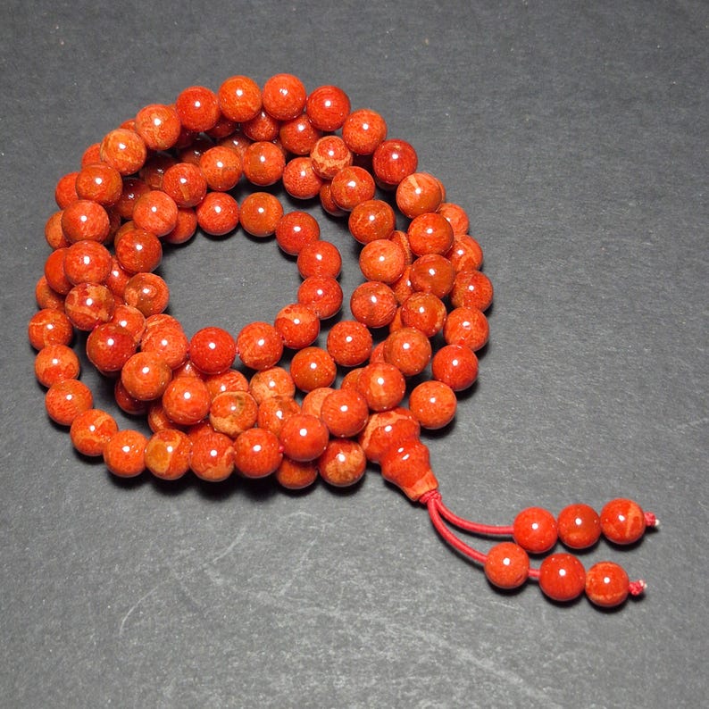May include: A strand of orange-red beads, likely coral, arranged in a circular pattern. The beads are round and have a slightly textured surface. A red tassel with additional beads hangs from the end. The background is a dark gray.
