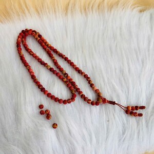Puede incluir: Un collar largo compuesto por peque&ntilde;as cuentas redondas y texturizadas en tonos rojos, naranjas y amarillos. Las cuentas est&aacute;n ensartadas en un cord&oacute;n fino y el collar termina en una borla anudada con varias cuentas sueltas cerca.