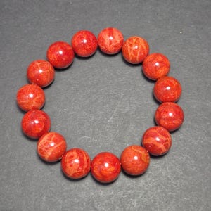 May include: A red coral bead bracelet. The round beads are a vibrant red color with natural patterns. The bracelet is displayed on a dark gray surface.