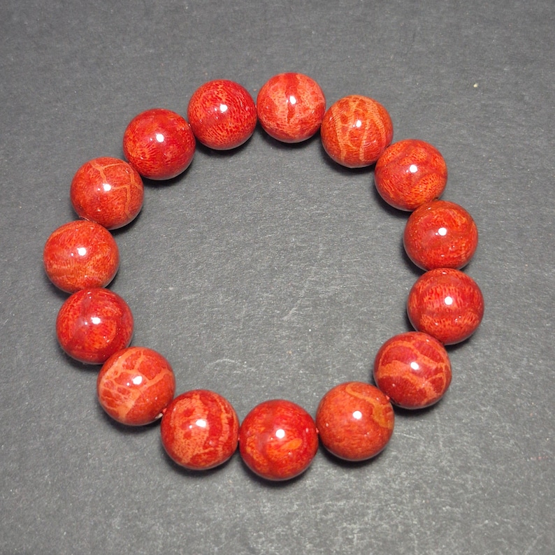 May include: A red coral bead bracelet. The round beads are a vibrant red color with natural patterns. The bracelet is displayed on a dark gray surface.