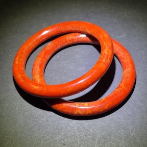 May include: Two orange bangles with a marbled appearance are displayed against a dark background. The bracelets are circular, with a smooth, glossy finish. The color is a vibrant, reddish-orange, with subtle variations in tone.