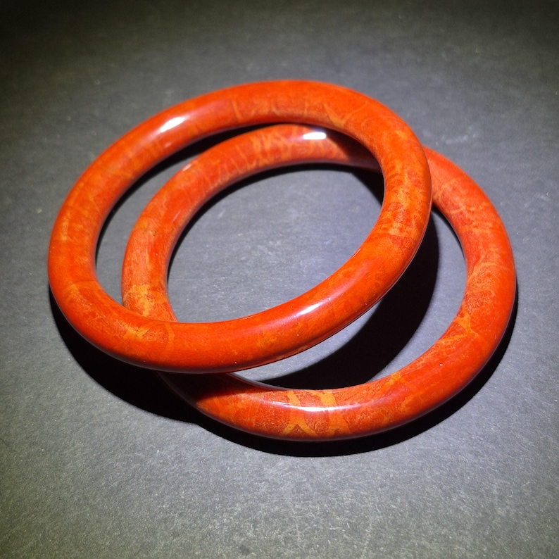 May include: Two orange bangles with a marbled appearance are displayed against a dark background. The bracelets are circular, with a smooth, glossy finish. The color is a vibrant, reddish-orange, with subtle variations in tone.