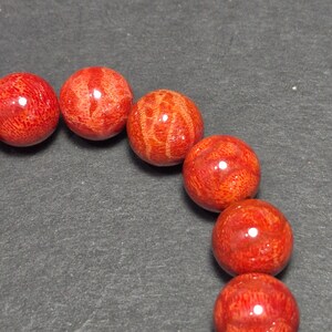 May include: Close-up of a strand of red coral beads. The beads are round, with a polished, glossy finish. The coral has a natural, textured pattern, and the color is a vibrant, deep red. The beads are strung together, creating a necklace or bracelet.