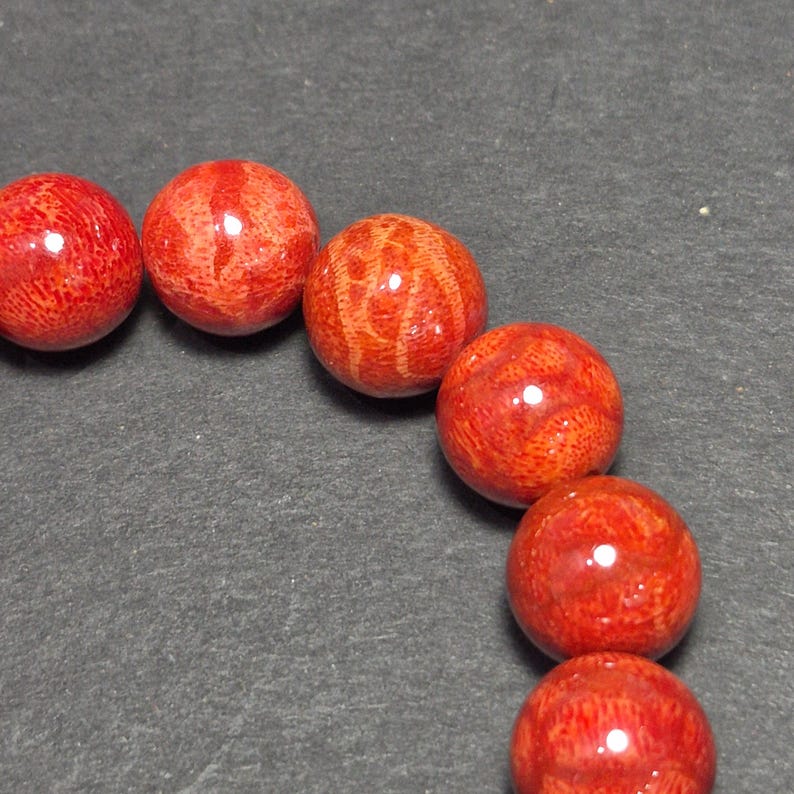 May include: Close-up of a strand of red coral beads. The beads are round, with a polished, glossy finish. The coral has a natural, textured pattern, and the color is a vibrant, deep red. The beads are strung together, creating a necklace or bracelet.