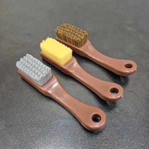Könnte beinhalten: Drei Schuhbürsten mit braunen Griffen und unterschiedlich farbigen Borsten: grau, gelb und braun. Jede Bürste hat einen Griff mit einem Loch zum Aufhängen. Die Borsten sind rechteckig und dicht gepackt.