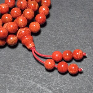 May include: Close-up of a strand of red coral beads. The beads are round and have a polished finish. A red tassel is attached to the end of the strand. The beads are a vibrant shade of red.