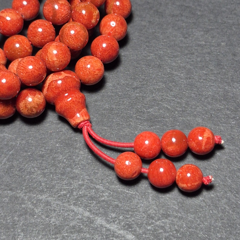 May include: Close-up of a strand of red coral beads. The beads are round and have a polished finish. A red tassel is attached to the end of the strand. The beads are a vibrant shade of red.