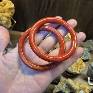 May include: Two red coral bangles with a natural, textured pattern. The circular bracelets are displayed on a hand, showcasing their vibrant color and unique design. These accessories are a striking addition to any jewelry collection.