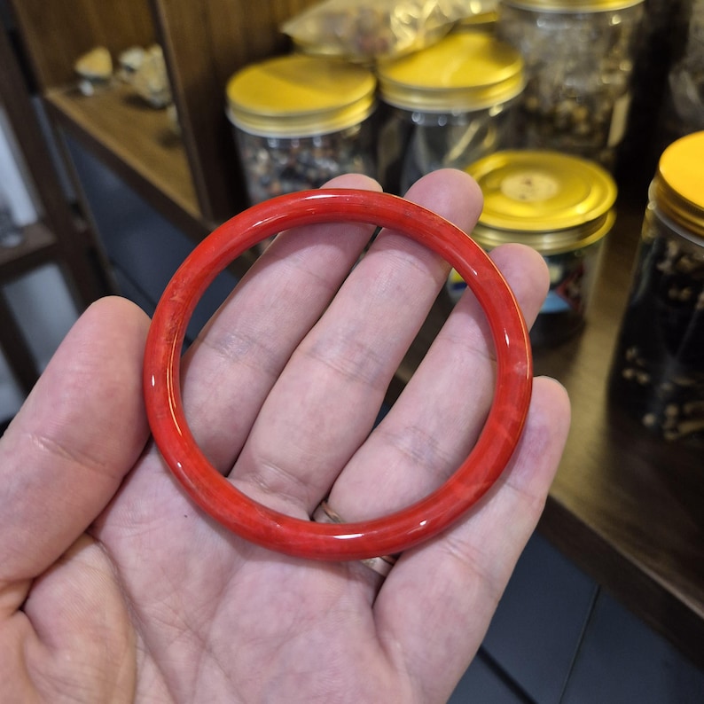 May include: A vibrant red bangle bracelet. The circular bracelet is a solid, glossy red with subtle variations in color. The bracelet is held in a hand, with jars and shelves in the background.