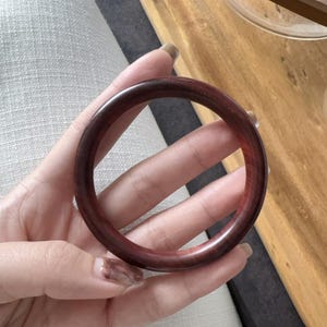 Pode incluir: Uma pulseira de madeira circular e polida, em um tom castanho-avermelhado profundo. A pulseira é lisa e parece ser feita de uma única peça de madeira. A pulseira é exibida em um fundo neutro, destacando sua textura e cor naturais.