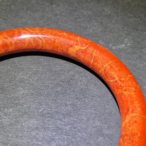 May include: Close-up of a vibrant orange bangle bracelet. The bracelet has a smooth, polished surface with intricate patterns in shades of orange and yellow. The jewelry is set against a dark background.