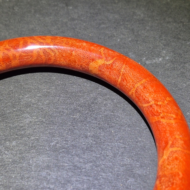 May include: Close-up of a vibrant orange bangle bracelet. The bracelet has a smooth, polished surface with intricate patterns in shades of orange and yellow. The jewelry is set against a dark background.