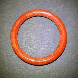 May include: A round, orange bangle bracelet with a mottled pattern. The bracelet is a solid band of color, with variations of orange and yellow throughout. The jewelry is set against a dark gray background.