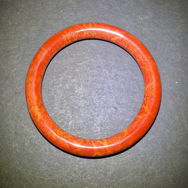 May include: A round, orange bangle bracelet with a mottled pattern. The bracelet is a solid band of color, with variations of orange and yellow throughout. The jewelry is set against a dark gray background.