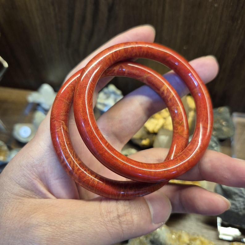 May include: Two red coral bangles with a polished finish. The bangles are a vibrant red color with natural patterns. The circular bracelets are held in a hand, showcasing their smooth texture and design.