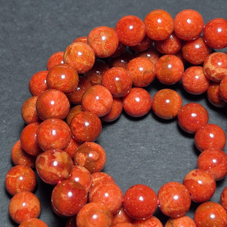 May include: Strand of round, reddish-orange beads. The beads have a polished, glossy finish and a mottled texture, with variations in color from light to dark orange. The beads are strung together, creating a necklace or bracelet.
