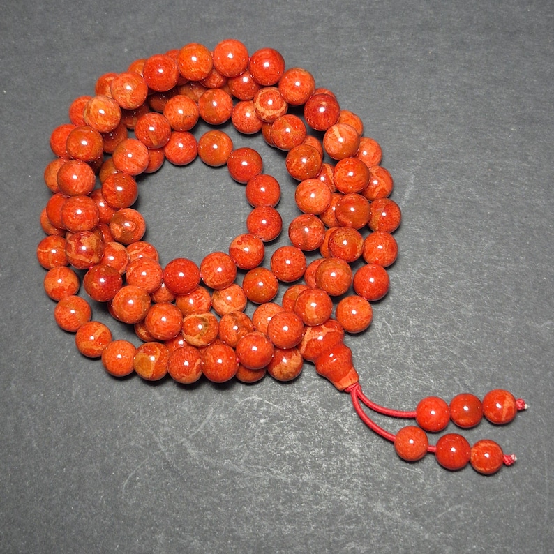 May include: A strand of orange-red beads, likely coral, arranged in a circular pattern. The beads are round and have a natural, textured appearance. A tassel of smaller beads and red string hangs from the strand. The beads are on a dark gray surface.