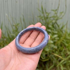 Puede incluir: Un brazalete circular liso hecho de una pieza sólida de piedra azul. El brazalete presenta un acabado pulido y brillante con variaciones sutiles de color, incluyendo motas de color azul claro y oscuro en todo el material.