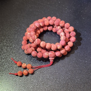 Puede incluir: Un collar circular de cuentas esf&eacute;ricas texturizadas en color rojo coral, dispuestas en espiral. Las cuentas tienen una superficie mate y porosa con un sutil veteado en rojo claro y rosa. Dos cordones rojos finos se extienden desde la pieza.