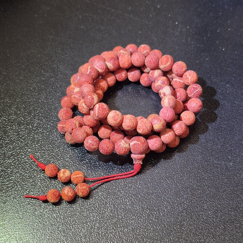 Puede incluir: Un collar circular de cuentas esf&eacute;ricas texturizadas en color rojo coral, dispuestas en espiral. Las cuentas tienen una superficie mate y porosa con un sutil veteado en rojo claro y rosa. Dos cordones rojos finos se extienden desde la pieza.