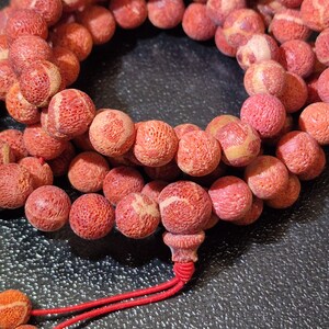 Puede incluir: Un collar de cuentas esf&eacute;ricas con una textura porosa similar al coral en tonos rojo anaranjado y crema. Las cuentas est&aacute;n ensartadas en un cord&oacute;n rojo fino, con una cuenta central m&aacute;s grande al final con una borla roja anudada.