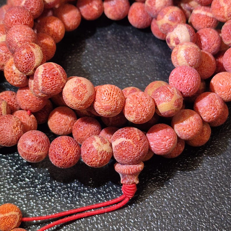 Puede incluir: Un collar de cuentas esf&eacute;ricas con una textura porosa similar al coral en tonos rojo anaranjado y crema. Las cuentas est&aacute;n ensartadas en un cord&oacute;n rojo fino, con una cuenta central m&aacute;s grande al final con una borla roja anudada.