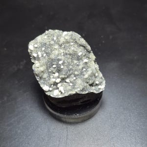 May include: A gray and white mineral specimen, possibly a druzy quartz, sits on a black, round display stand. The mineral has a textured surface with small, rounded crystals. The background is a dark gray.