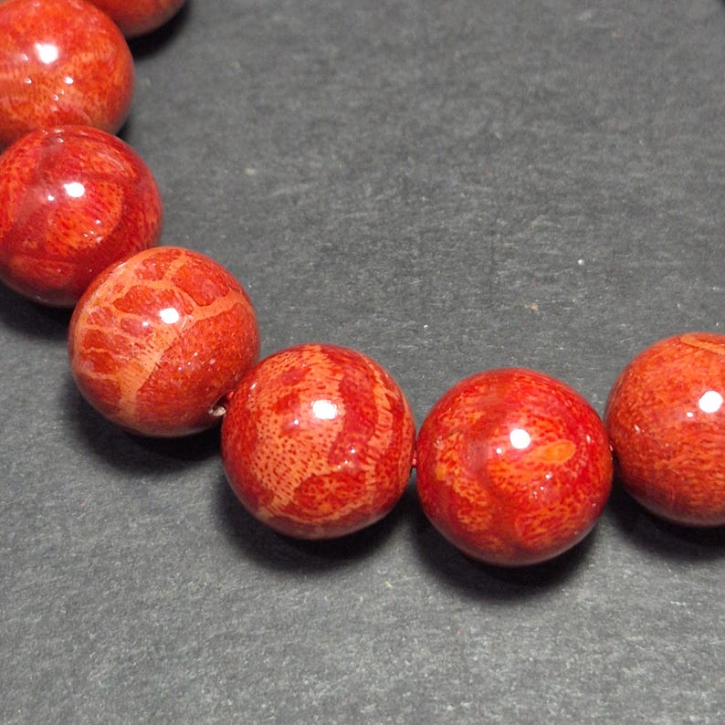 May include: Close-up of a strand of red coral beads. The beads are round, with a polished, glossy finish. The coral has a natural, textured pattern and is strung together. The background is a dark gray surface.