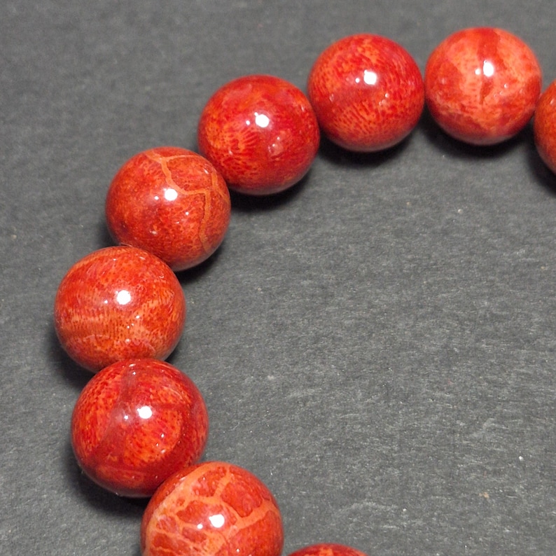 May include: Close-up of a bracelet featuring round, polished red coral beads. The beads have a natural, textured surface with variations in color and pattern. The bracelet is displayed against a dark gray background, highlighting the vibrant red color of the coral.