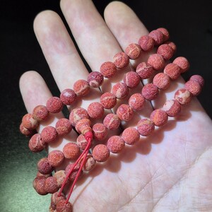 Puede incluir: Un collar de cuentas redondas y porosas de color rojo anaranjado con texturas naturales visibles y patrones de vetas claras. Las cuentas est&aacute;n ensartadas en un cord&oacute;n rojo fino, formando un bucle continuo dispuesto en varias capas.