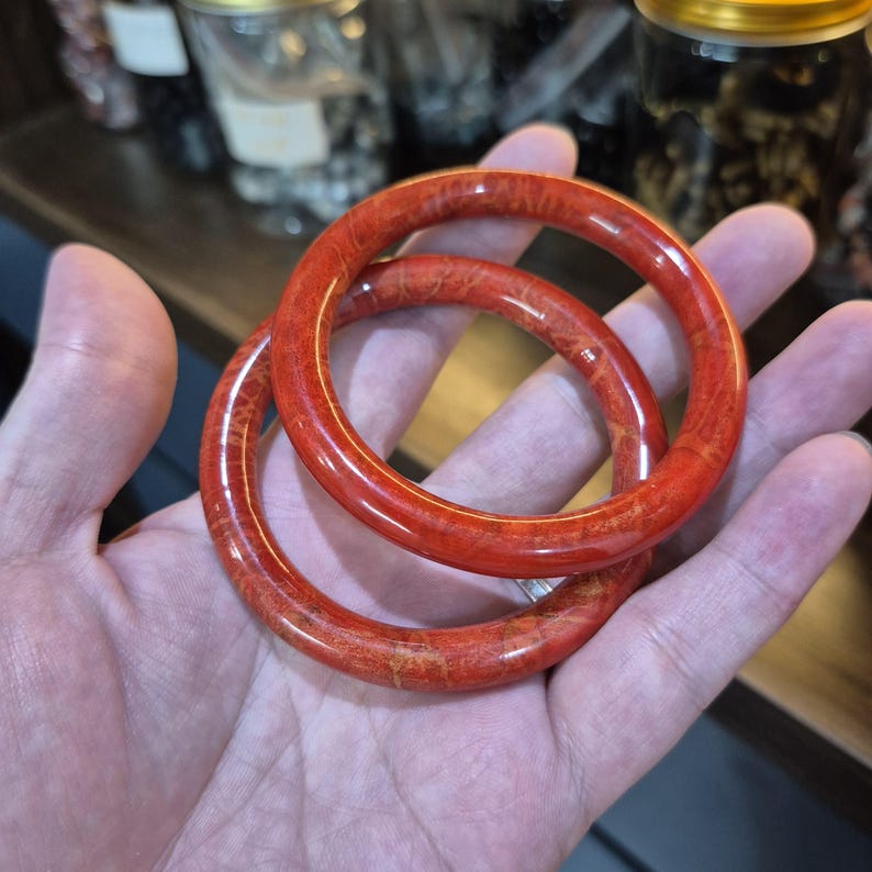 May include: Two polished, red bangles with a natural wood grain pattern are held in a hand. The bangles are circular and appear to be made of a reddish-orange material. The background is blurred, suggesting a shop setting.