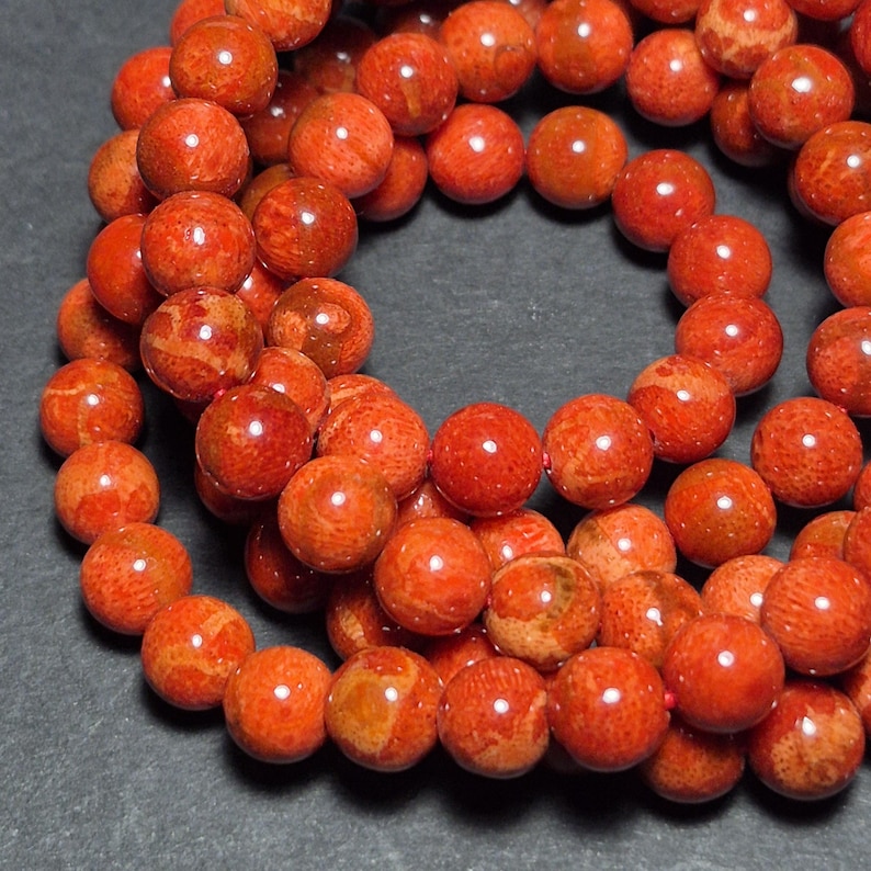 May include: A strand of round, polished coral beads in shades of red and orange. The beads have a natural, textured appearance with variations in color and pattern. The beads are strung together, creating a necklace or bracelet.