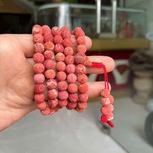 Puede incluir: Un collar de cuentas redondas, porosas y de color coral con una superficie texturizada. Las cuentas est&aacute;n ensartadas en m&uacute;ltiples bucles, conectadas por un cord&oacute;n rojo brillante que se extiende con un peque&ntilde;o grupo de cuentas a juego.