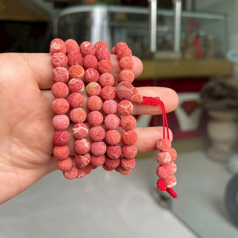 Puede incluir: Un collar de cuentas redondas, porosas y de color coral con una superficie texturizada. Las cuentas est&aacute;n ensartadas en m&uacute;ltiples bucles, conectadas por un cord&oacute;n rojo brillante que se extiende con un peque&ntilde;o grupo de cuentas a juego.