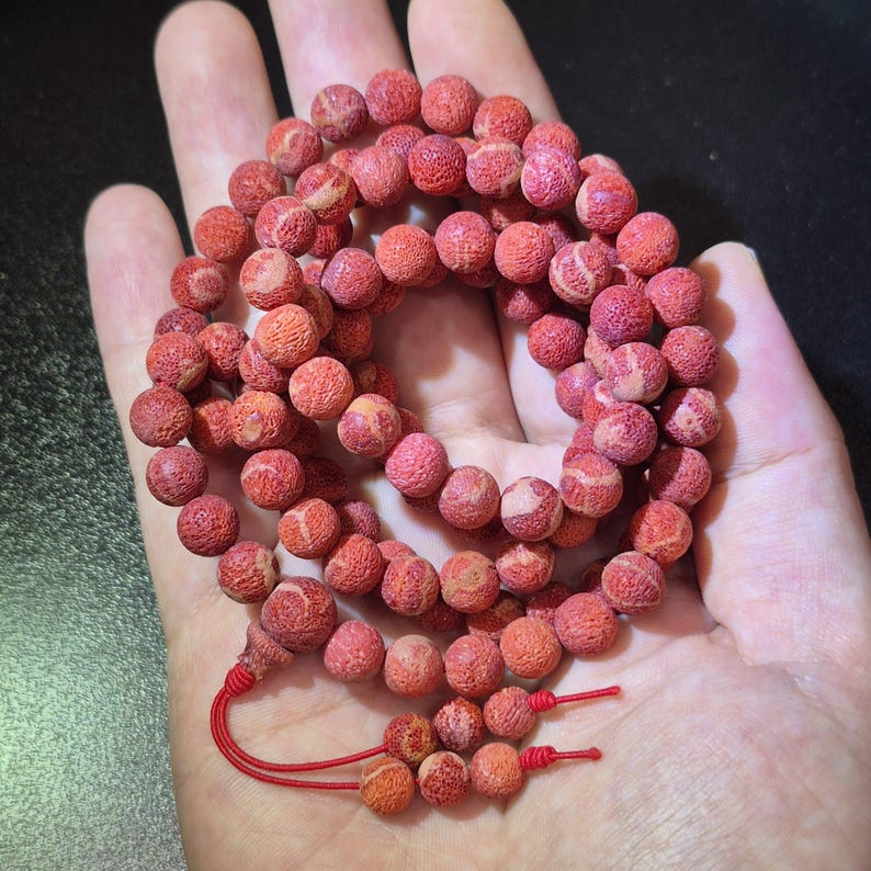 Puede incluir: Un collar de cuentas esf&eacute;ricas, porosas y de color rojo con una superficie texturizada. Las cuentas est&aacute;n ensartadas en forma circular, con un cord&oacute;n rojo que se extiende desde el extremo y termina en dos peque&ntilde;as cuentas colgantes.