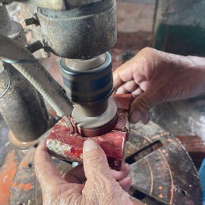 May include: A close-up view of a person using a machine to grind a red rectangular object. The machine has a rotating grinding wheel. The object is held in place by the person's hands. The machine and object are in a workshop setting.