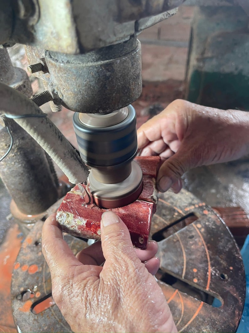 May include: A close-up view of a person using a machine to grind a red rectangular object. The machine has a rotating grinding wheel. The object is held in place by the person's hands. The machine and object are in a workshop setting.