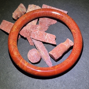 May include: A round, reddish-orange coral bracelet with a polished finish. The bracelet is surrounded by pieces of coral in varying shapes and sizes, displaying a porous texture. The background is a dark gray surface.