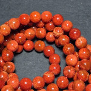 May include: A strand of round, orange-red beads. The beads have a natural, textured appearance with variations in color and pattern. The beads are strung together, creating a necklace or bracelet.