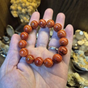 May include: A beaded bracelet featuring round, reddish-orange beads with natural wood grain patterns. The bracelet is held in a hand, showcasing the polished, spherical beads. The bracelet is a jewelry item.