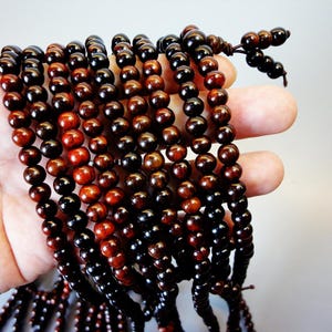 May include: Several strands of wooden beads in varying shades of brown and black are held in a hand. The beads are round and strung together, with a darker knot at the end of each strand. The beads have a polished finish.