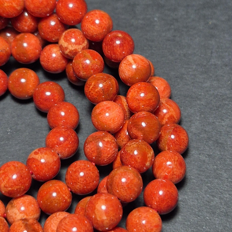 May include: Close-up of a strand of round, orange-red coral beads. The beads have a polished, glossy finish and are arranged in a curved pattern against a dark gray background. The beads are approximately 8mm in diameter.