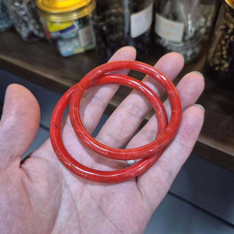 May include: Two bright red bangles, likely made of coral or a similar material, are displayed. The bangles are circular and have a smooth, polished surface. The image is taken from a close-up angle, highlighting the color and texture of the jewelry.