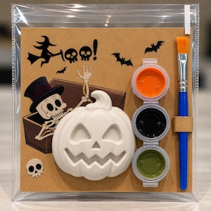 May include: A Halloween craft kit featuring a white ceramic pumpkin, three small paint pots in orange, black, and green, and a blue paintbrush. The cardboard backing displays a skeleton in a top hat, bats, and the text Boo! in black ink.