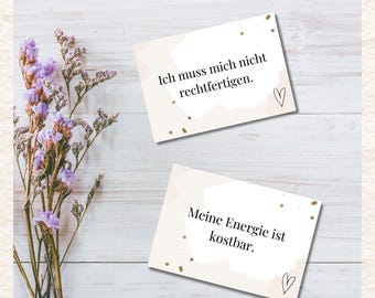 May include: Two beige cards with black text and gold accents. The top card reads "Ich muss mich nicht rechtfertigen." The bottom card reads "Meine Energie ist kostbar." A sprig of purple flowers is on the left.