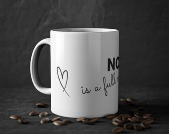May include: White ceramic mug with a black handle, featuring a black heart and the phrase "NO is a full sentence." Coffee beans surround the mug. Steam rises from the mug against a dark background.