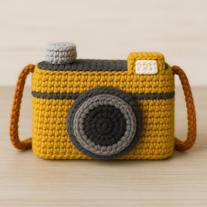 May include: A handcrafted crochet camera in yellow, grey, and brown. The camera has a large lens, a top button, and a strap. The body is yellow with grey accents and a brown strap. The camera is made with soft yarn.