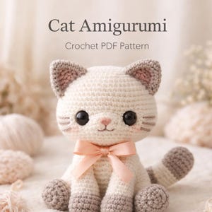 Op de afbeelding: Een gehaakte kat amigurumi met een crèmekleurig lichaam, grijze oren, poten en staart. De kat heeft grote zwarte ogen, roze wangen en een roze strik. De tekst "Cat Amigurumi Crochet PDF Pattern" staat bovenaan.