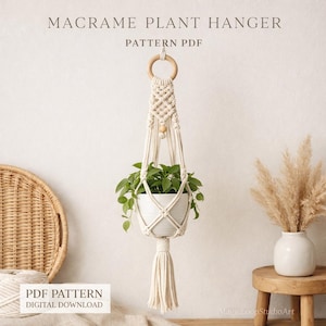 Puede incluir: Colgador de plantas de macramé color crema con un anillo y una cuenta de madera, que sostiene una maceta blanca con plantas verdes. El diseño incluye una borla en la parte inferior. El texto en la imagen dice "MACRAME PLANT HANGER PATTERN PDF" y "PDF PATTERN DIGITAL DOWNLOAD".